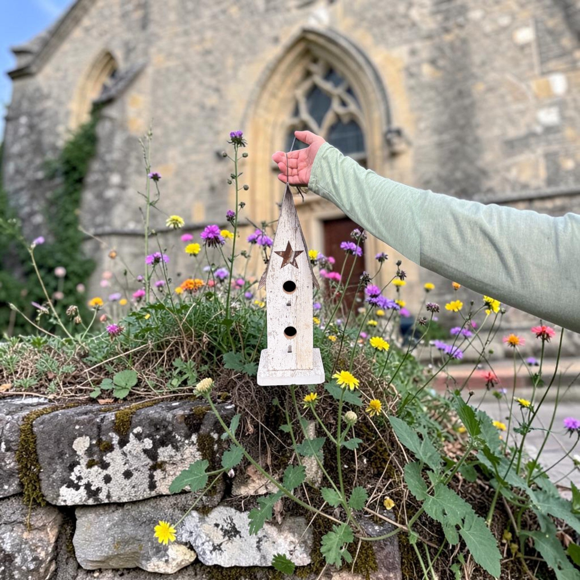 Person holding a wooden birdhouse in front of a stone building with flowers and plants.